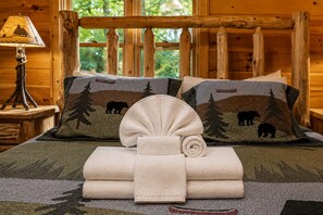 A wooden cabin room with a bed featuring a forest-themed pillow and a rolled towel. - A Touch of Bearadise - Mountain View | Pet Friendly | Close to Town! | Game Room | Firepit | (Morganton)