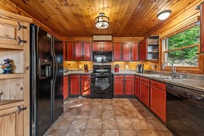 A kitchen with wooden cabinets, a refrigerator, a stove, and a window overlooking greenery. - A Touch of Bearadise - Mountain View | Pet Friendly | Close to Town! | Game Room | Firepit | (Morganton)