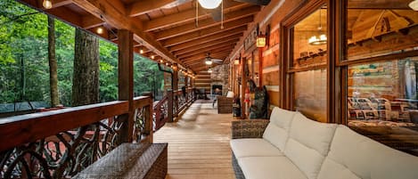 A covered porch with a wooden ceiling, a white sofa, wicker furniture, and a view of the forest.