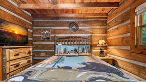 A log cabin interior with a bed, a television, and a bear-themed artwork. - Bear Claw Lodge - Pet Friendly | Firepit | Outdoor Fireplace | Hot Tub (Cherry Log)