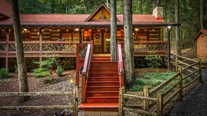 A cabin with a staircase leading to a wooden deck, surrounded by trees. - Bear Claw Lodge - Pet Friendly | Firepit | Outdoor Fireplace | Hot Tub (Cherry Log)