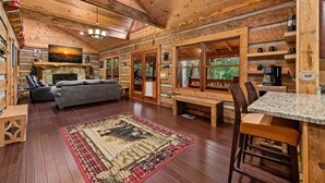 A log cabin interior with a fireplace, seating area, dining space, and large windows. - Bear Claw Lodge - Pet Friendly | Firepit | Outdoor Fireplace | Hot Tub (Cherry Log)
