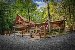 A rustic cabin with a porch and a covered deck, surrounded by a forest. - Bear Claw Lodge - Pet Friendly | Firepit | Outdoor Fireplace | Hot Tub (Cherry Log)