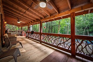 A covered outdoor area with wooden flooring, wicker furniture, and a view of a forest. - Bear Claw Lodge - Pet Friendly | Firepit | Outdoor Fireplace | Hot Tub (Cherry Log)