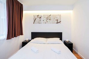 A hotel room with a bed, two bedside tables, a window with curtains, and three wall-mounted artworks.
