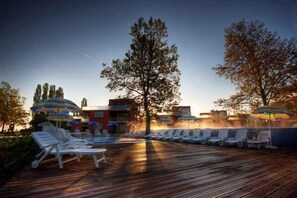Eine Holzterrasse mit Liegestühlen und Sonnenschirmen, ein Schwimmbecken und ein Gebäude mit roter Fassade.