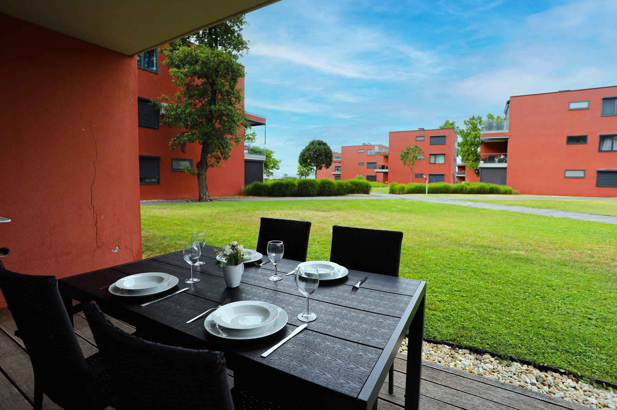 Une table à manger installée à l’extérieur, offrant une vue sur des bâtiments rouges et une pelouse verdoyante.