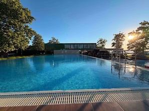 Ein klares Schwimmbecken mit Holzterrasse im Vordergrund, einem überdachten Sitzbereich und einem Gebäude mit großen Fenstern im Hintergrund.