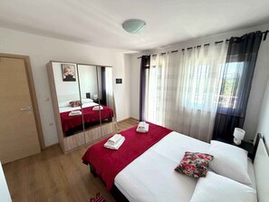 Hotellihuone, jossa on sänky, vaatekaappi, peili, yöpöytä ja ikkuna, jossa on verhot.