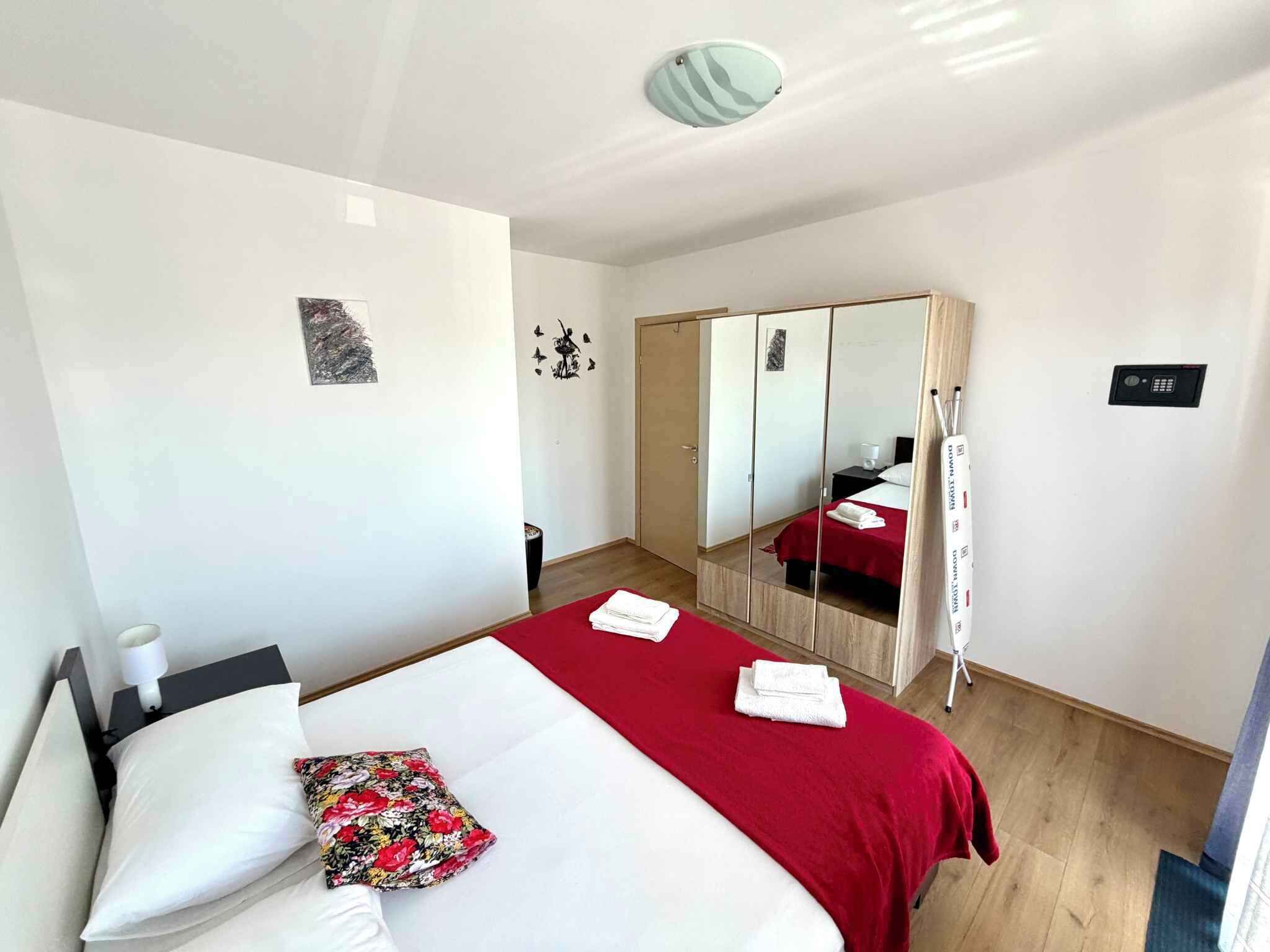 A hotel room with a bed, a red bedspread, a floral pillow, a bedside table with a lamp, a mirror, and a wardrobe.