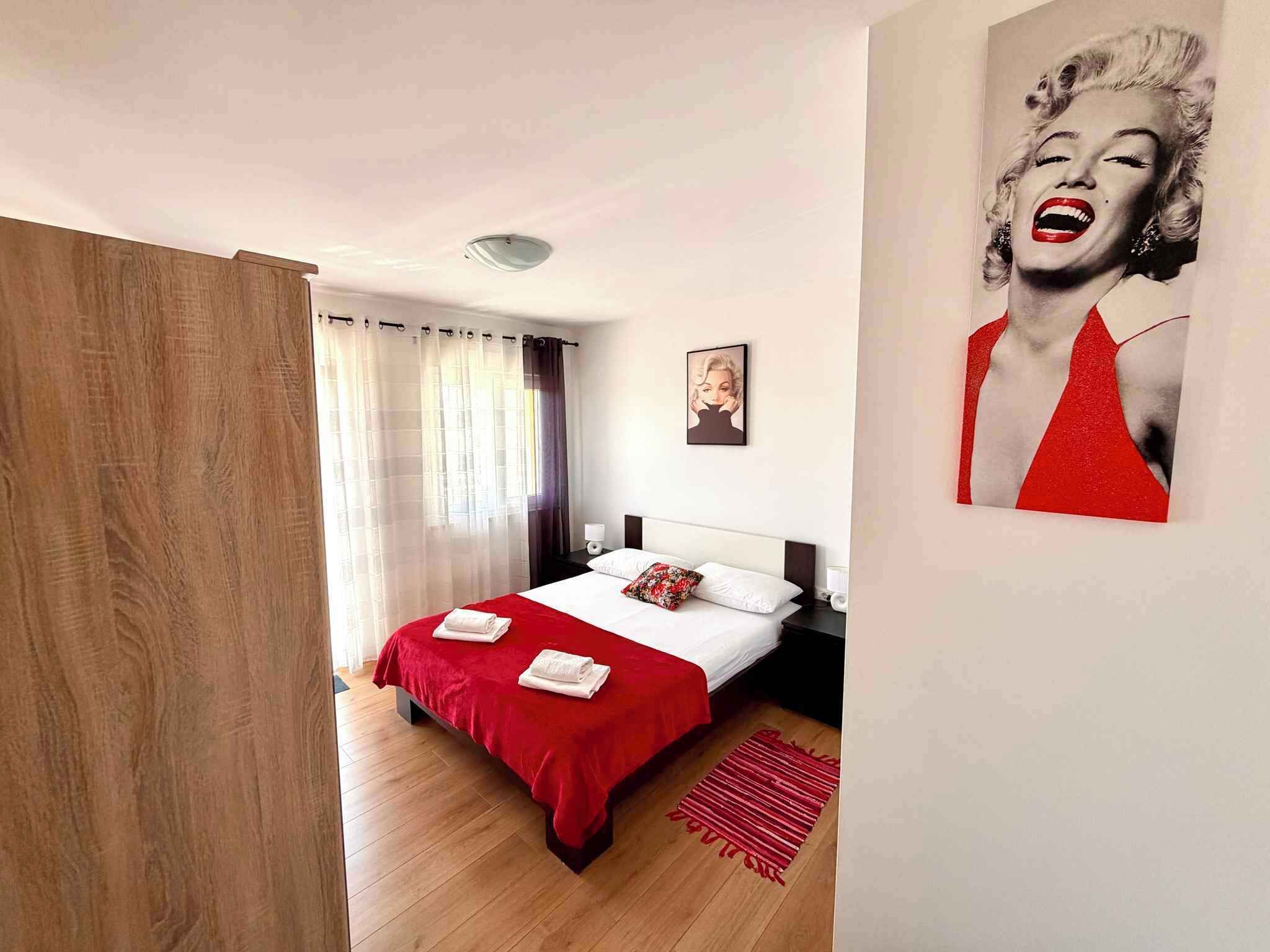 Une chambre à coucher avec un lit, des rideaux, une table de chevet, une lampe, un tableau encadré et un tapis rouge.