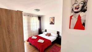 Ein Schlafzimmer mit einem Bett, Vorhängen, einem Nachttisch, einer Lampe, einem gerahmten Bild und einem roten Teppich.