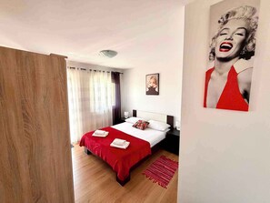 Ein Schlafzimmer mit einem Bett, Vorhängen, einem Nachttisch, einer Lampe, einem gerahmten Bild und einem roten Teppich.