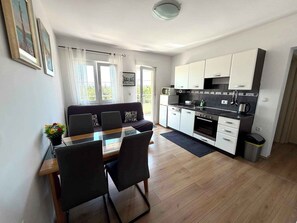 Eine moderne Küche mit weißen Schränken, ein Essbereich mit einem Glastisch und eine Sitzecke mit einem Sofa.