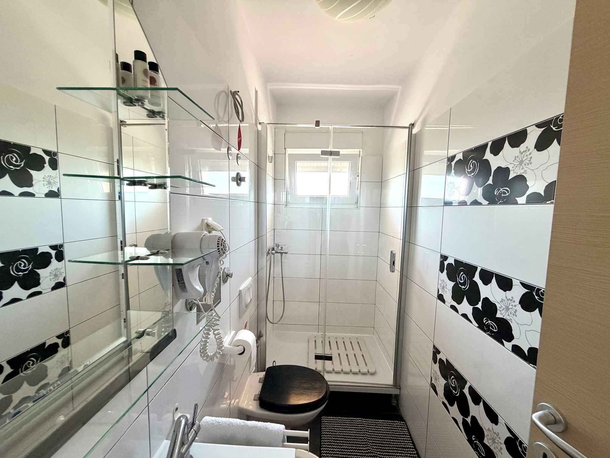 Ein schmales Badezimmer mit Duschkabine aus Glas, Toilette und Waschbecken mit einer Glasablage darüber.
