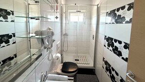 Ein schmales Badezimmer mit Duschkabine aus Glas, Toilette und Waschbecken mit einer Glasablage darüber.