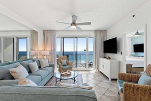 Ein geräumiges Wohnzimmer mit einer Couch, Sesseln, einem Couchtisch und einem Fernseher. Ein Balkon bietet Blick auf das Meer.
