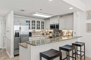 Eine moderne Küche mit Edelstahl-Kühlschrank, Mikrowelle, Wasserspender und einer Arbeitsplatte aus Granit. Weiße Schränke und ein Barhocker sind vorhanden.