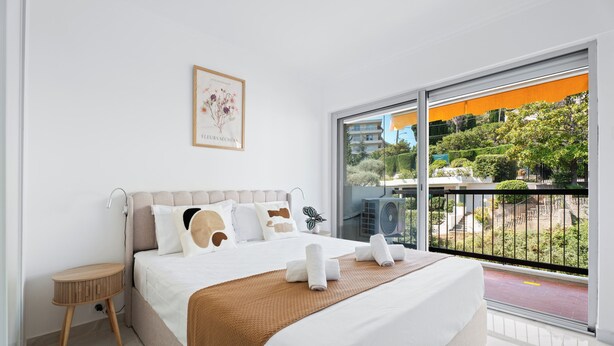 Ein Hotelzimmer mit einem Bett, einem kleinen Nachttisch, einem großen Fenster mit Blick auf Grünflächen und Gebäude sowie einem gerahmten Bild an der Wand.