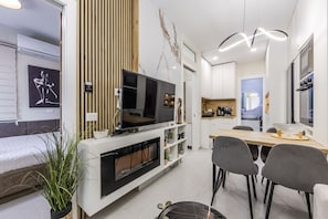 Ein modernes Wohnzimmer mit Flachbildfernseher, einem Essbereich mit Holztisch und Stühlen sowie einer Küche im Hintergrund.