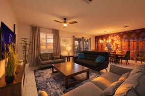 A living room with a sofa, a coffee table, and a dining area with a chandelier. - 9 Bedroom Themed Villa Sleeps 24 with Private Pool Spa and Arcade (Kissimmee)