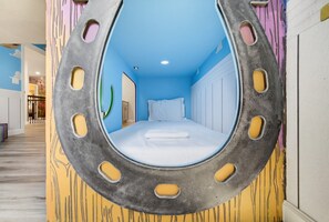 Une pièce avec un lit placé au centre d’une structure circulaire ressemblant à une fenêtre, une porte sur la gauche et un couloir menant à une autre pièce.