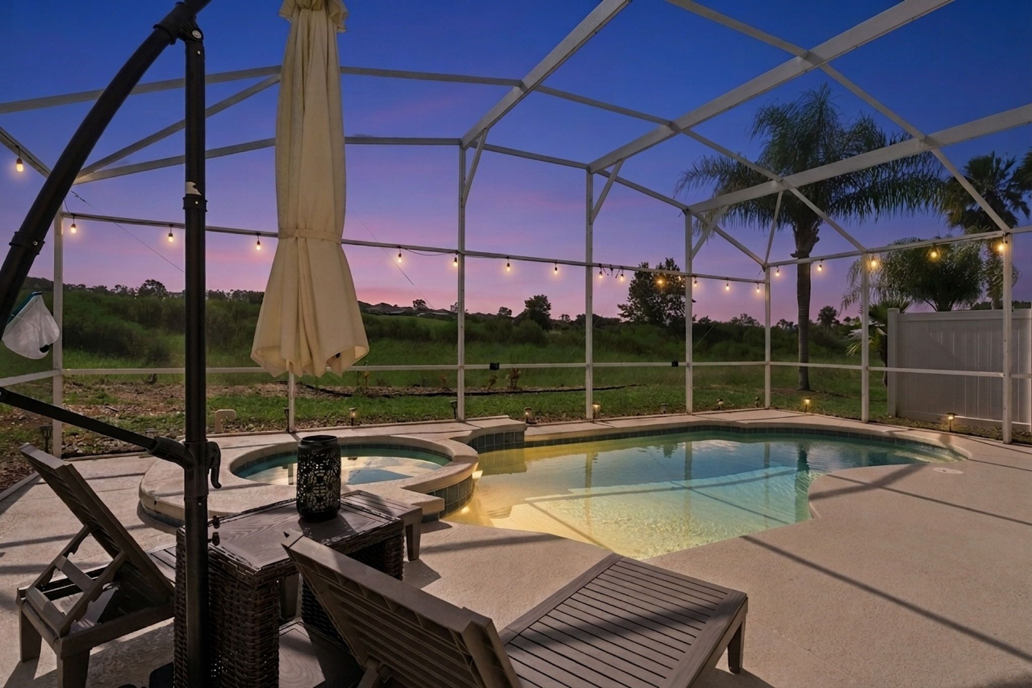 A covered outdoor swimming pool with lounge chairs and a table, surrounded by a white fence and string lights.