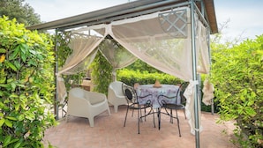 Un área de estar al aire libre, techada, con una mesa y sillas, rodeada de vegetación.