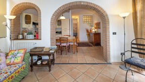Una sala de estar acogedora con un sofá, una mesa de centro y una zona de comedor con sillas y una mesa. Se observan arcos de ladrillo que conectan las habitaciones, y se ve una cocina al fondo.
