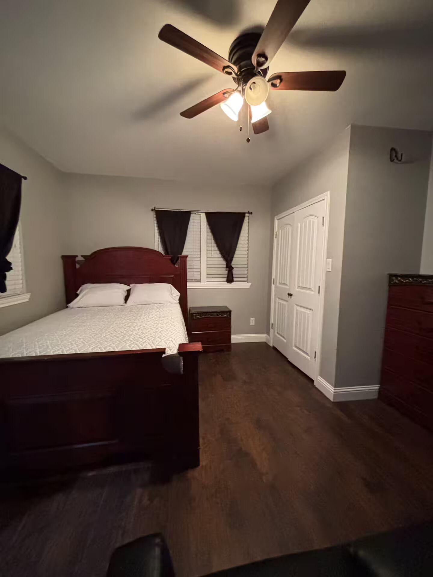 Quarto com cama de madeira, ventoinha no teto, mesa de cabeceira, cômoda e uma porta.