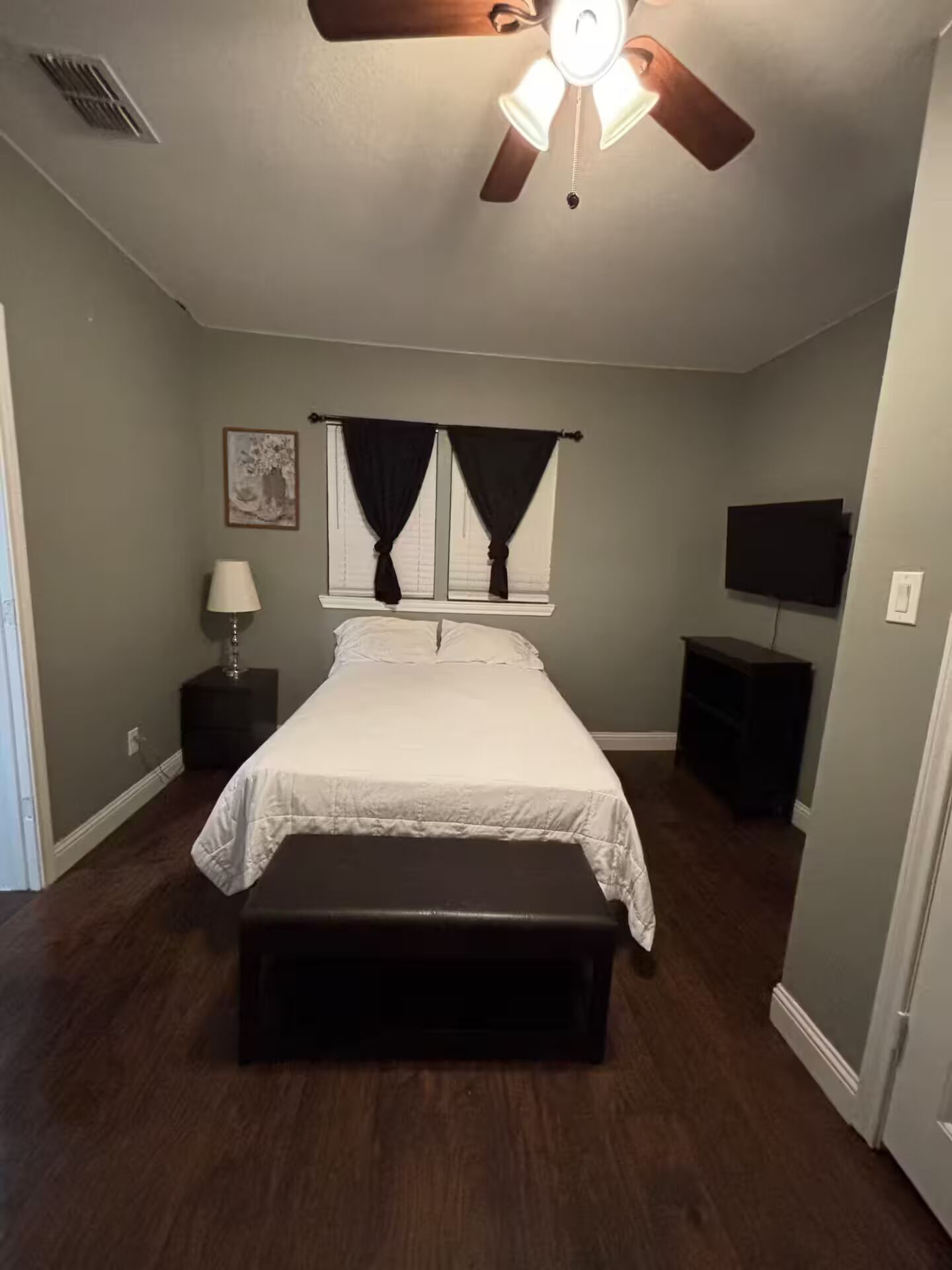 A bedroom with a bed, a ceiling fan, a nightstand, a lamp, a TV, and a bench.