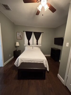 A bedroom with a bed, a ceiling fan, a nightstand, a lamp, a TV, and a bench.