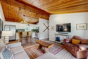 Uma sala de estar ampla com teto de madeira, uma cozinha ao fundo, um sofá de couro, uma televisão e uma área de jantar com cadeiras.