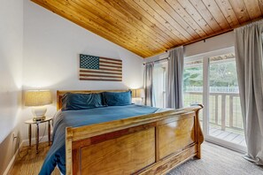A bedroom with a wooden bed, a nightstand with a lamp, a view of the outdoors through a window with curtains, and an American flag on the wall.