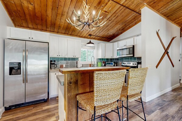 A kitchen with a wooden ceiling, stainless steel appliances, a bar area with woven chairs, and a wall-mounted X-shaped decoration.