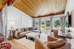 A spacious living area with a high wooden ceiling, a sofa set, a coffee table, a TV, and a view of the outdoors. - Great Location:  Bike, Walk, Run & Ski (Stowe)