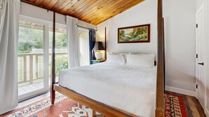 Un dormitorio con techo de madera, cama con ropa de cama blanca, un cuadro en la pared y vistas al exterior por la ventana.