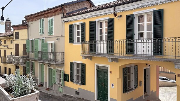 Uma fileira de edifícios amarelos com venezianas verdes, uma varanda com grades de ferro forjado e um vaso de planta em primeiro plano.