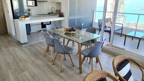 A modern kitchen with a refrigerator, sink, and dining area with chairs and a glass table. - Rbk Amplio Dpto Familiar con linda vista (Concón)