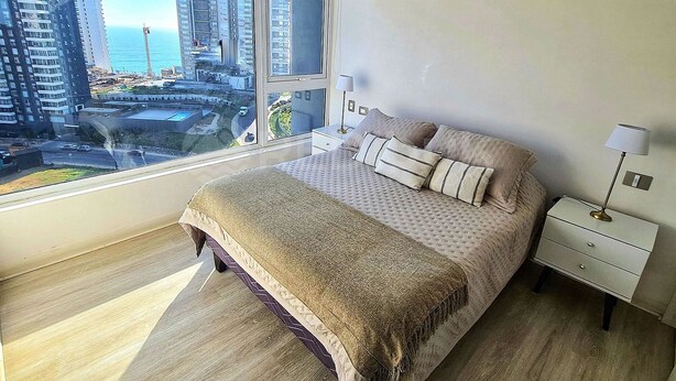 A bedroom with a large window, a bed with pillows, a bedside table with a lamp, and a view of tall buildings. - Rbk Amplio Dpto Familiar con linda vista (Concón)