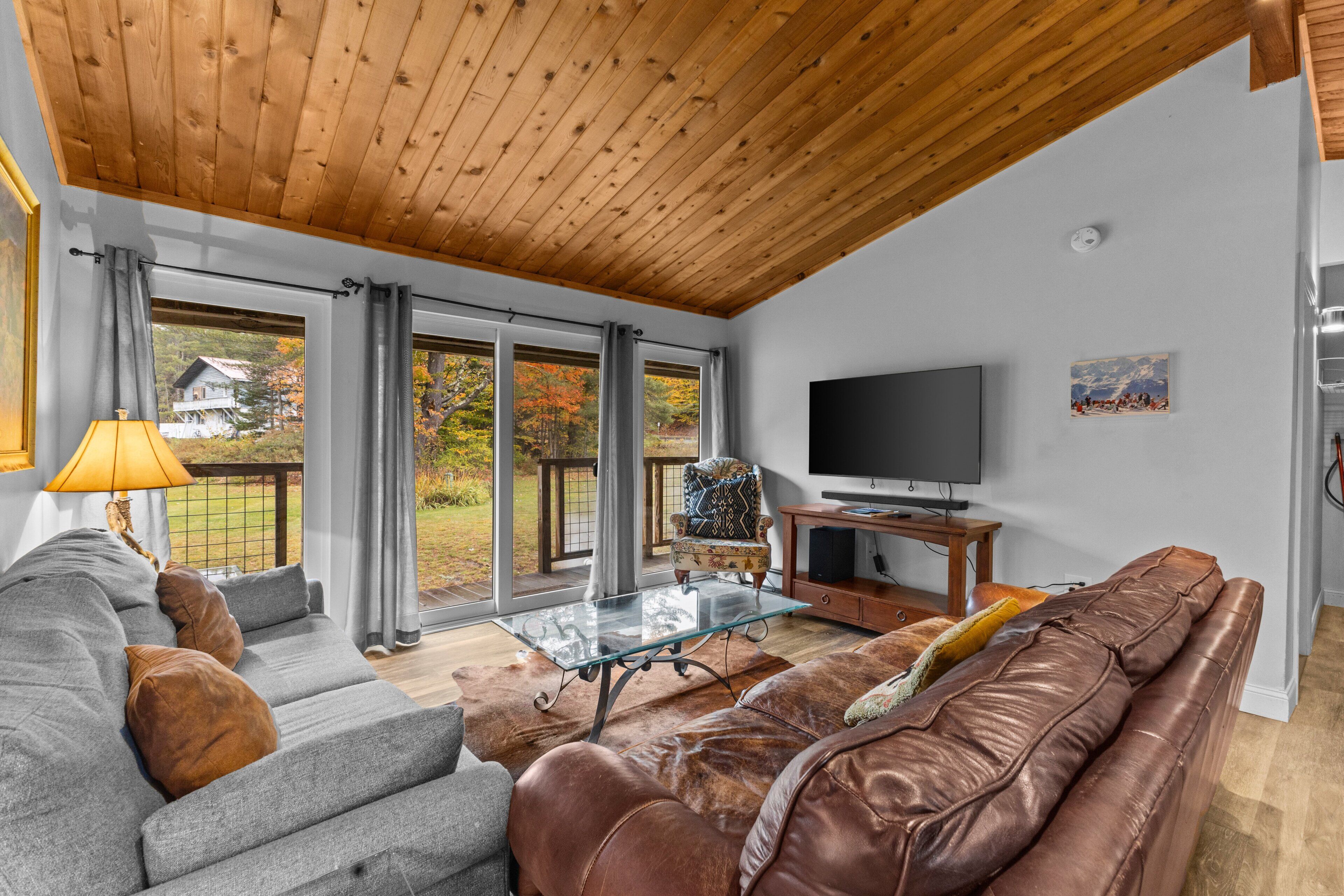 Un salon moderne avec un plafond en bois, un canapé gris, un divan en cuir marron, une table basse en verre et un téléviseur.