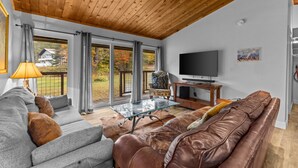 Un salon moderne avec un plafond en bois, un canapé gris, un divan en cuir marron, une table basse en verre et un téléviseur.