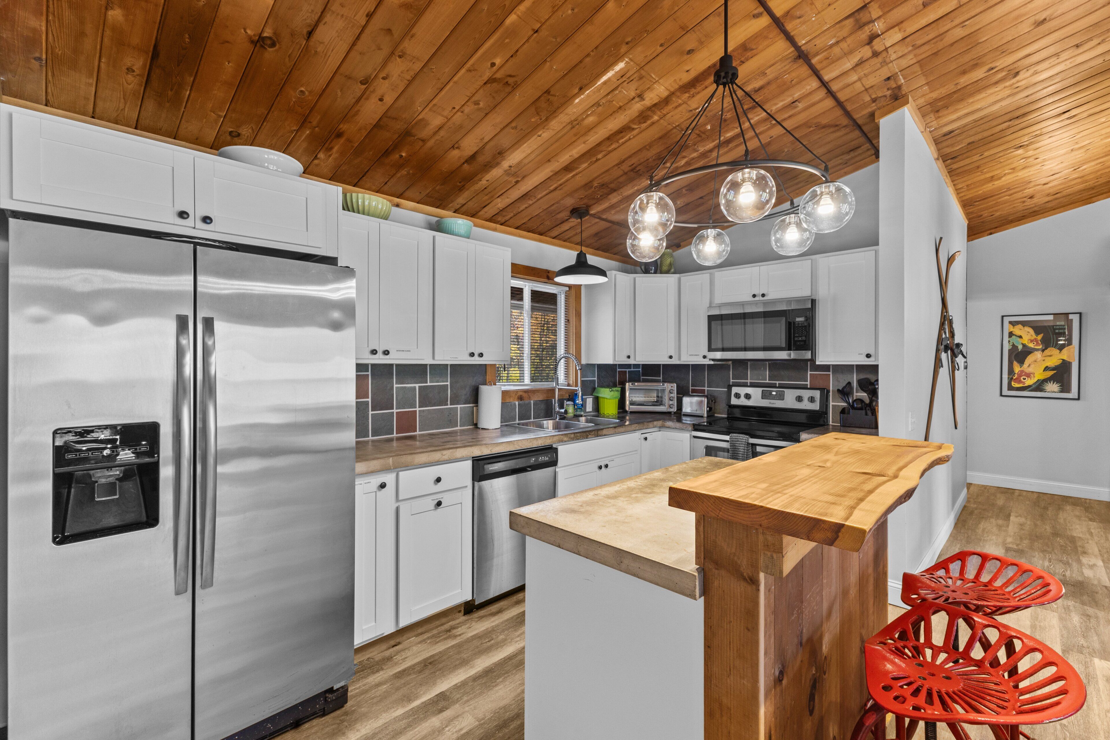 A modern kitchen with stainless steel appliances, white cabinetry, a wooden ceiling, and a central island with a bar stool.
