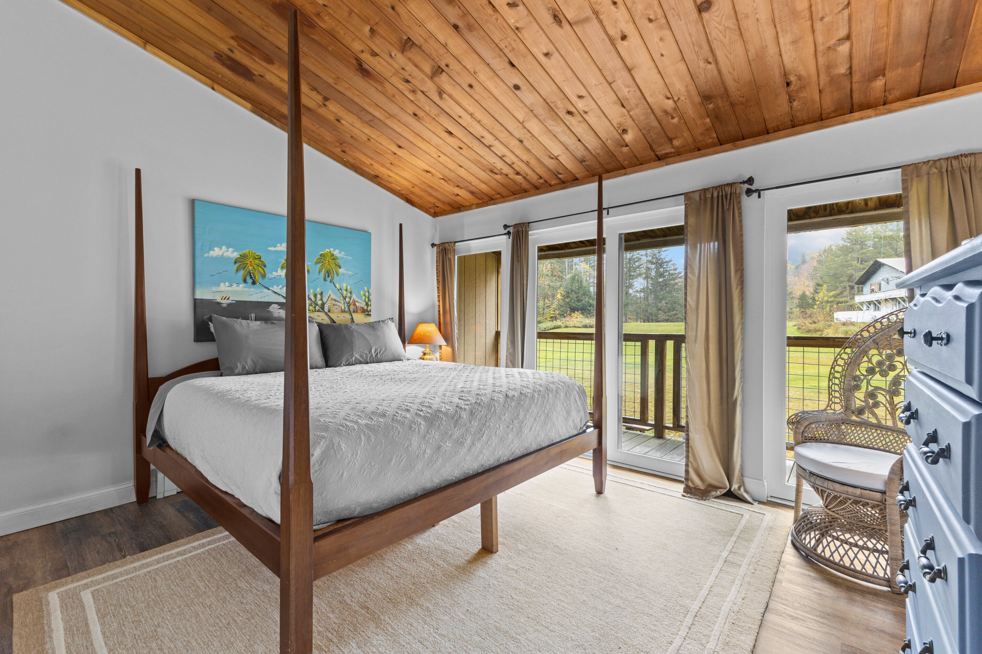 Une chambre à coucher avec un lit à baldaquin, un tableau, une lampe, un fauteuil en osier et une vue sur l’extérieur.