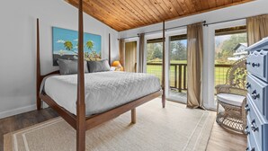 Une chambre à coucher avec un lit à baldaquin, un tableau, une lampe, un fauteuil en osier et une vue sur l’extérieur.