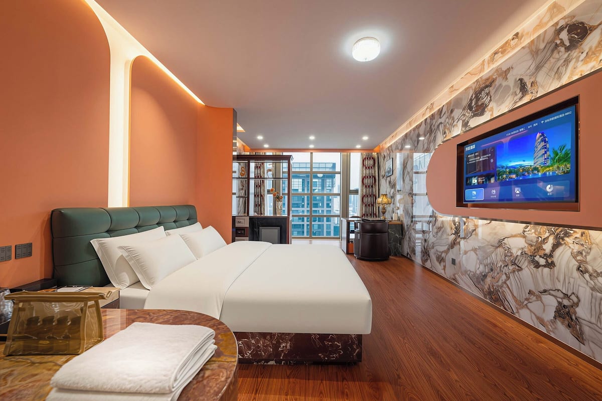 A modern hotel room with a large bed, a TV mounted on the wall, and a view of the cityscape through the window.