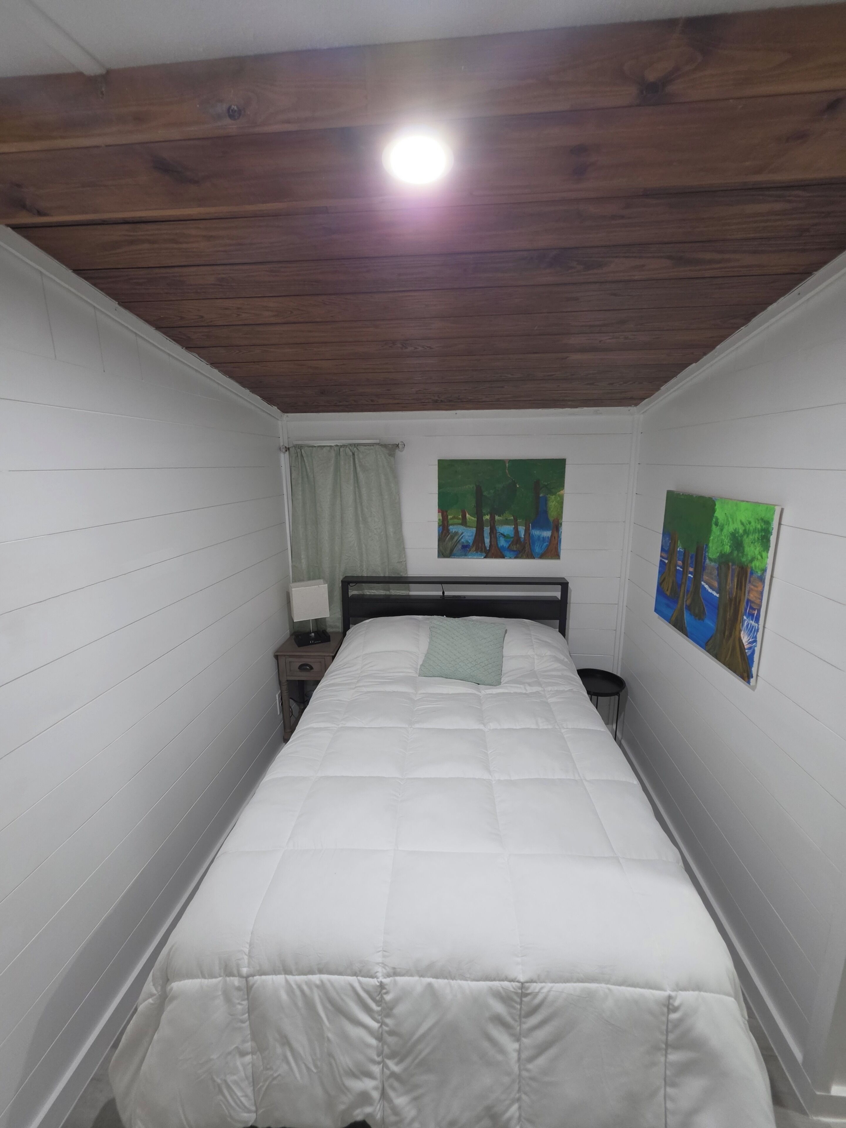 Une chambre avec un plafond en bois, un lit avec une literie blanche, une table de chevet, une lampe et deux tableaux accrochés au mur.