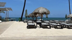 A beach with lounge chairs and thatched umbrellas, a wooden deck, and a clear view of the ocean. - Beach Condo 2 Pools Walk to Beach Bars Eats (Punta Cana)