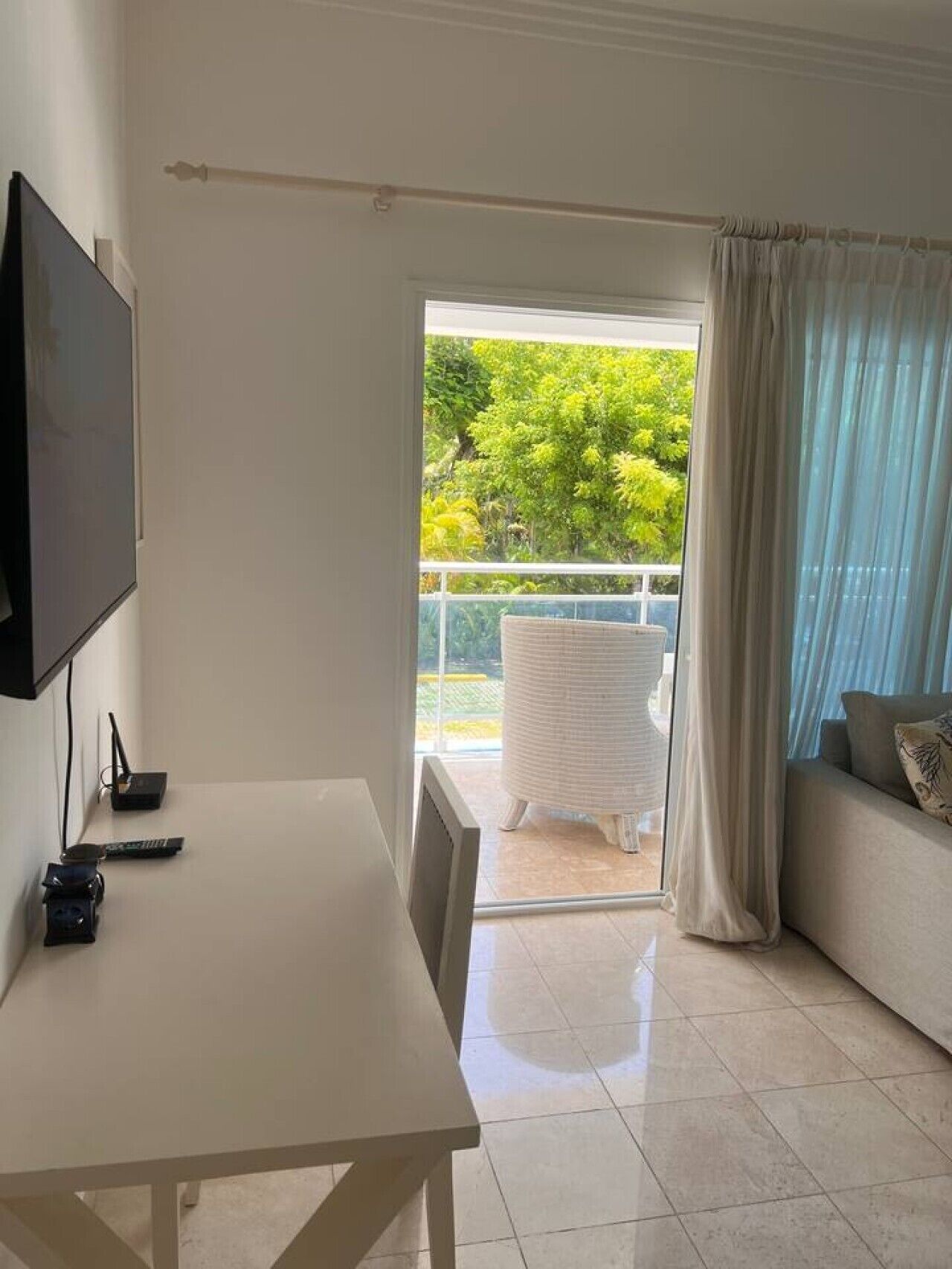 Quarto com televisão, mesa, sofá e varanda com vista para o verde.