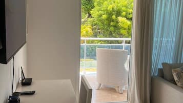 Quarto com televisão, mesa, sofá e varanda com vista para o verde.